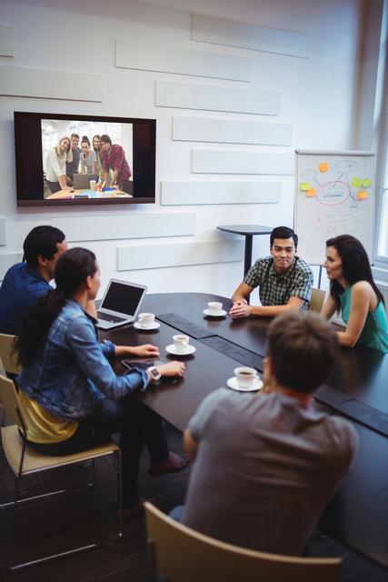 A group of business executives are gathered around a conference table, engaging in a video conference with a team displayed on the screen. Perfect for illustrating modern business communication, teamwork, and remote collaboration in corporate settings. Useful for business articles, remote work resources, and corporate training materials.