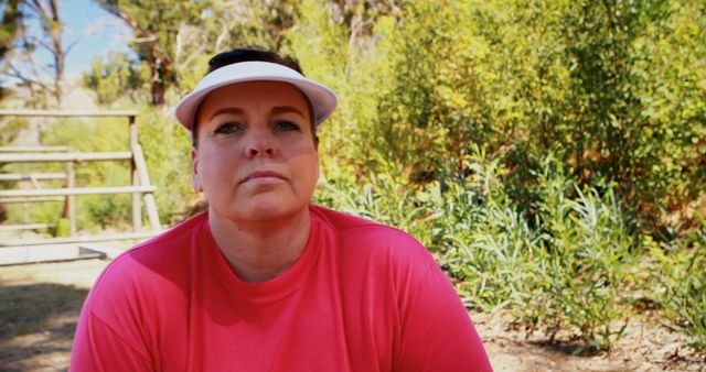 Middle-aged woman wearing a pink shirt and white visor, sitting outdoors amid a lush natural backdrop. Ideal for promoting outdoor relaxation, casual lifestyle, or nature-related products and activities.