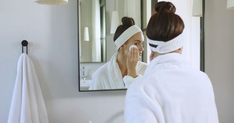 Woman Cleansing Face with Cotton Pad in Bathroom Mirror Reflection