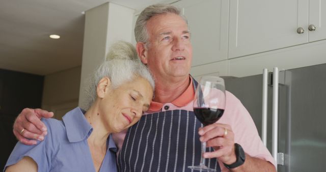 Happy caucasian senior couple wearing aprons embracing and drinking wine. active and healthy retirement lifestyle at home.