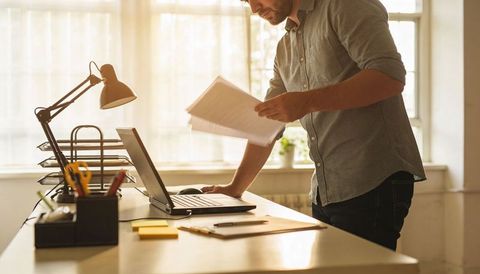 Man reviewing documents at sunlit desk with laptop, organized workspace and modern lamp