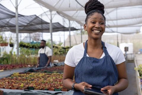 Smiling african american nursery worker using tablet while tending seedlings in greenhouse