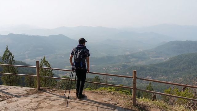 Standing hiker holding tripod at mountain overlook watching misty valley and forested hills