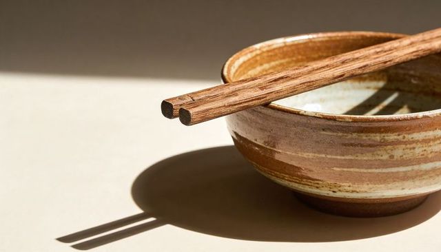 Stoneware bowl with wooden chopsticks casting long shadows over pale table minimal ceramic