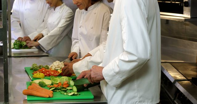 Chefs cutting various fresh vegetables together around a stainless steel table, using sharp knives and green chopping boards. Ideal for illustrating teamwork, food preparation, restaurant kitchens, culinary education, and professional cooking environments.