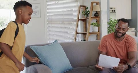 Dad reading school update while son returns home with backpack in bright living room