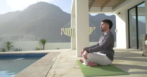 Indian man meditating on yoga mat by pool with mountain view on modern terrace