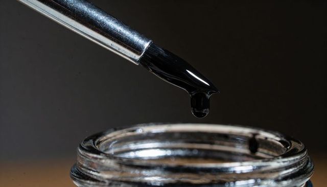 Fountain pen nib dropping glossy black ink drop above open glass ink bottle macro