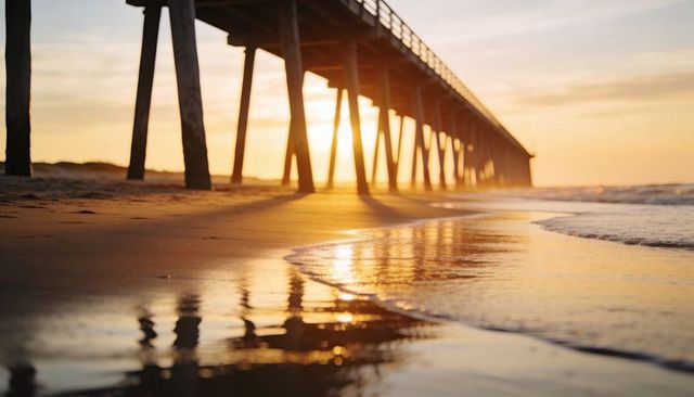 Sunrise pier casting golden reflection on wet sand, long wooden jetty leading to horizon