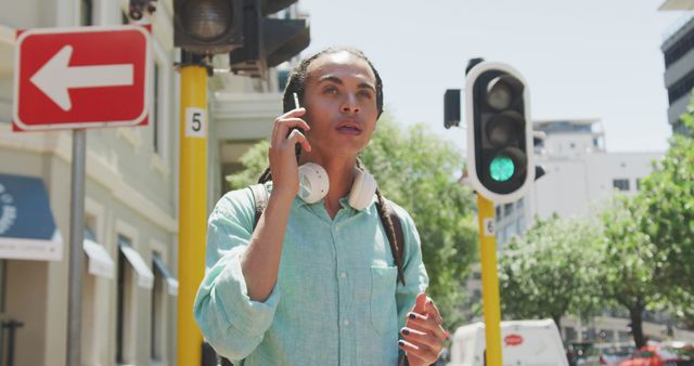 Young man with headphones around neck talking on smartphone while standing at busy city intersection near traffic lights. Suitable for themes of urban lifestyle, modern communication, public transport, technology use in daily life. Ideal for use in blogs, advertisements, or articles focusing on city living or telecommunications.