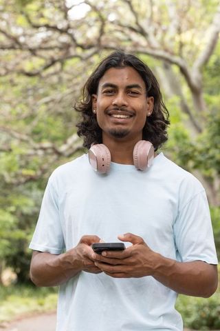 African american young man smiling in park wearing headphones and using smartphone
