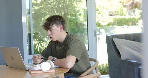 Teen male working from home at dining table using laptop and headphones with natural light