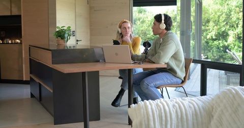 Young duo recording podcast at home in modern kitchen with laptop, microphones, headphones