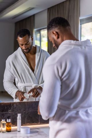 African american man applying face cream at bathroom vanity in white bathrobe