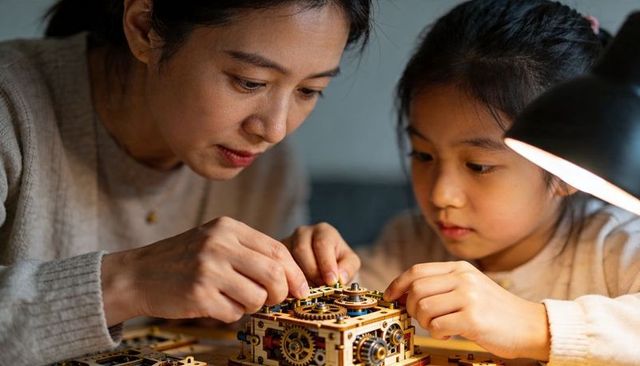 Mother guiding daughter assembling wooden mechanical gear model at table under lamp