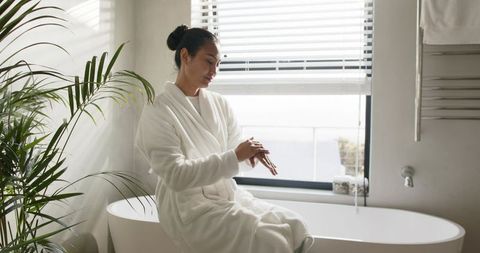 Mid-adult asian woman applying lotion while sitting on freestanding tub in sunlit bathroom