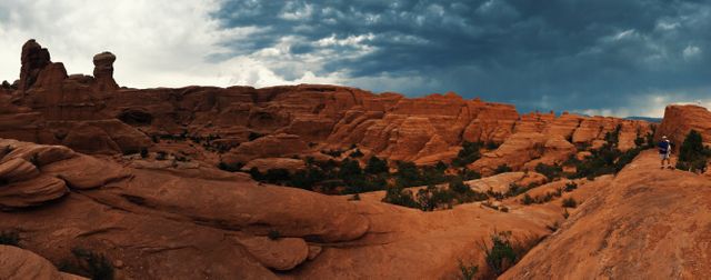 Two people hiking along large red rock formations under dark, ominous storm clouds. Image capturing raw natural beauty, perfect for promoting outdoor activities, adventure tourism, and nature photography. Could be used in travel blogs, magazines, or advertisements for hiking gear and outdoor adventure companies.