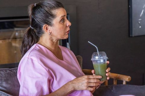 Mature woman in pink scrubs drinking green juice with reusable straw on sofa