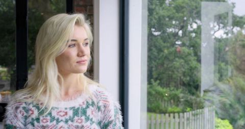 Blonde woman standing by floor-to-ceiling window gazing at garden in fuzzy pastel sweater