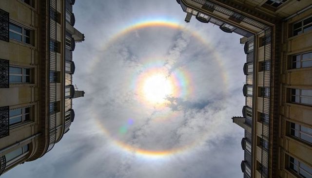 Circular sun halo and vivid corona framing upward view between stone balconies, city canyon