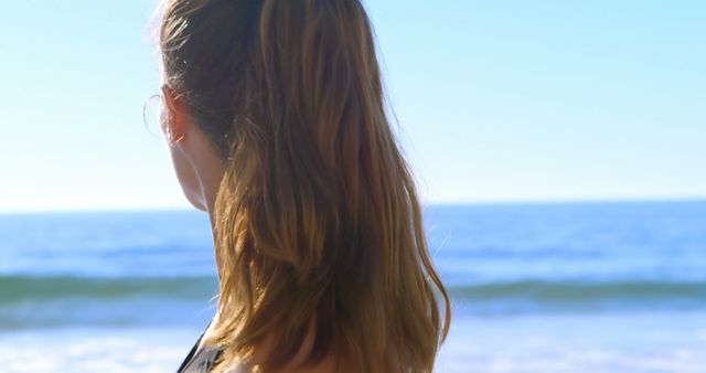 Woman with long hair standing on the beach looking out at the ocean on a sunny day. Ideal for promoting travel, relaxation, spa, mental health, and recreation. Can be used for advertisements, travel blogs, wellness articles, and social media content.
