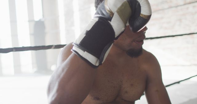 Boxer wearing gloves taking a moment to catch breath in the boxing ring. Ideal for topics on training, fitness, sports dedication, men’s health, athletic endurance, gym promotional material, strength conditioning.