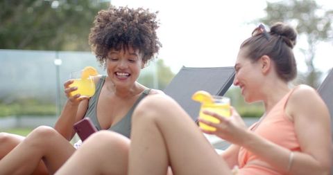 Female friends relaxing on pool deck holding citrus cocktails and checking smartphone