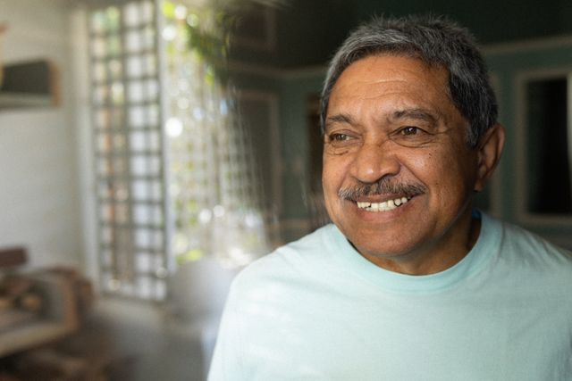 Senior African American man standing by a window in a bedroom, smiling warmly. Ideal for use in articles or advertisements about retirement, senior living, self-isolation during the COVID-19 pandemic, and promoting positive lifestyle choices for older adults.