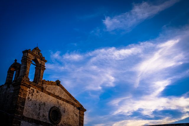 Ancient building with rustic bell tower stands against a dramatic evening sky filled with vivid clouds, evoking tranquility and historical grandeur. Ideal for use in travel blogs, historical articles, serene mood portrayals, and vintage architectural studies.