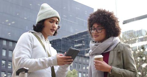 Diverse female friends checking smartphone on city sidewalk with suitcase and coffee