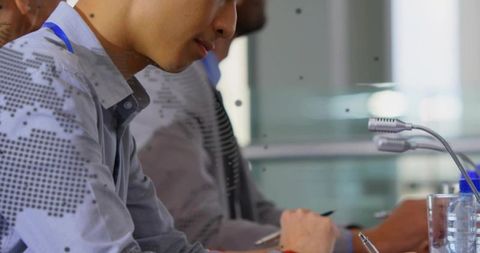 Asian professional taking notes at conference table with microphones and blue lanyard