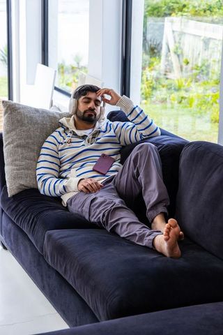 Indian man reclining on navy sofa wearing headphones and striped hoodie relaxing indoors