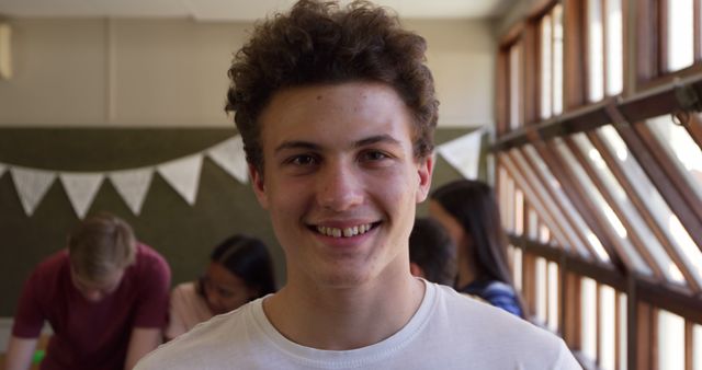 Young man with curly hair smiling, standing indoors with bunting and natural light from windows in background. Group of friends being socially active. Suitable for use in materials about youth, community events, social gatherings, and cheerful atmospheres.