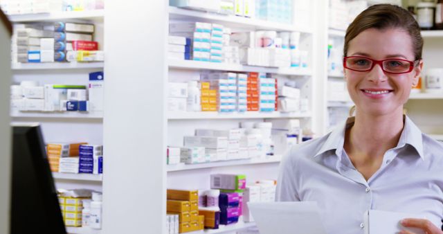 Photo shows a female pharmacist wearing red glasses, smiling while standing in front of shelves filled with medication boxes. This image can be used for healthcare-related articles, pharmacy promotions, or educational materials on medication management and pharmacy services.