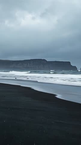 Vertical video of stormy black-sand beach with basalt cliffs, rolling swell and white foam