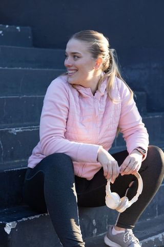 Smiling young woman in pink jacket holding headphones sitting on outdoor steps