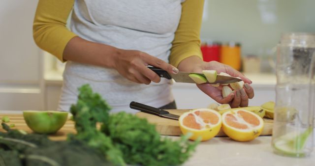 Mid section of biracial woman preparing healthy drink, cutting fruit and vegetables in kitchen. domestic life, spending quality free time relaxing at home.