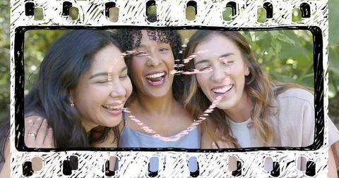 Three women laughing together in sunlit park with filmstrip frame