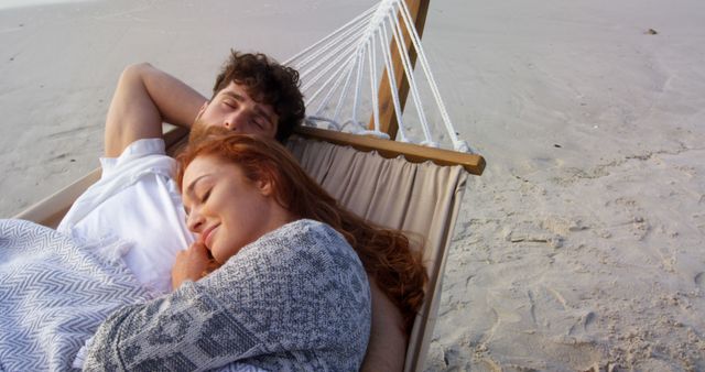 A young Caucasian couple enjoys a peaceful nap together in a hammock on a sandy beach, with copy space. Their relaxed posture and serene environment suggest a moment of tranquility and romantic connection.