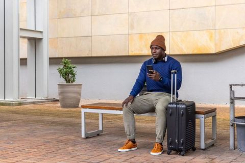 African american traveler waiting on bench with suitcase and smartphone in urban plaza