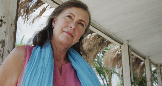 Middle-aged woman, wearing pink blouse and blue scarf, standing under gazebo, looking thoughtfully into distance. Suitable for concepts related to aging, reflection, peace, contemplation, and summer relaxation or vacation.