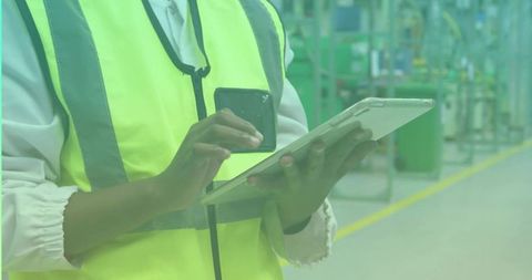 Technician operating tablet and smartphone on factory floor in high-visibility vest