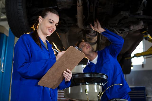 Mechanics preparing a check list while his colleague examining a car engine in the background