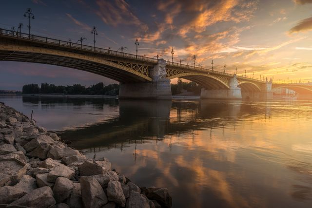 Depicts a steel bridge spanning a calm river during sunrise with a beautiful sky and serene reflections on the water. Ideal for use in travel brochures, city travel blogs, architecture and landmark articles. Can be used to highlight scenic sunrise views and historic landmarks.