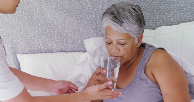 Elderly woman receiving assistance from a young caregiver as she drinks water in bed. Ideal for marketing home healthcare services, caregiving guides, nursing assistance materials, and articles on elderly well-being.