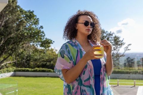 Curly-haired woman sipping fresh orange juice on sunny terrace overlooking city skyline