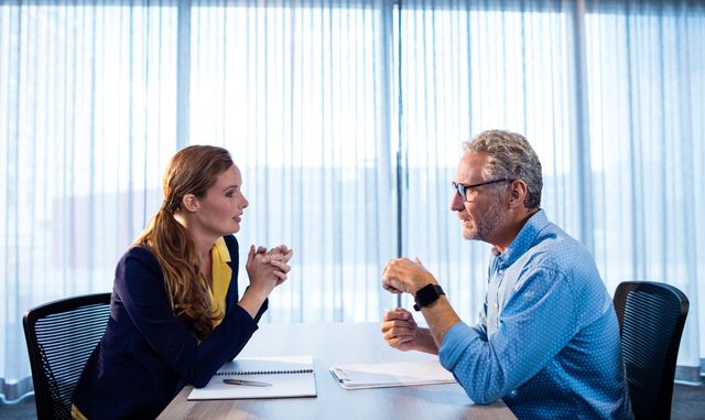 Two business colleagues are engaged in a discussion at an office table. They are focused and appear to be planning or strategizing. This image can be used to depict professional interactions, teamwork, and corporate meetings in various business-related contexts.