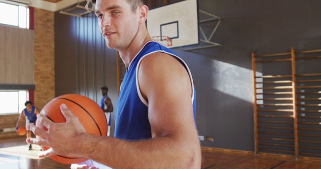 A young man holding a basketball on an indoor court, with teammates in the background. Could be used for promoting sports activities, fitness programs, team building, or educational content focused on physical education and youth sports.
