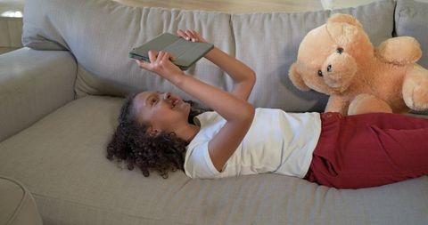 Smiling child lying on couch holding tablet overhead with teddy bear in cozy sunlit living room
