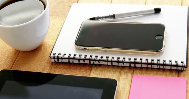 A smartphone lies on a spiral notebook next to a pen, with a cup of coffee and tablet nearby, with copy space. These items suggest a work or study environment, indicating a break or a moment of organization.